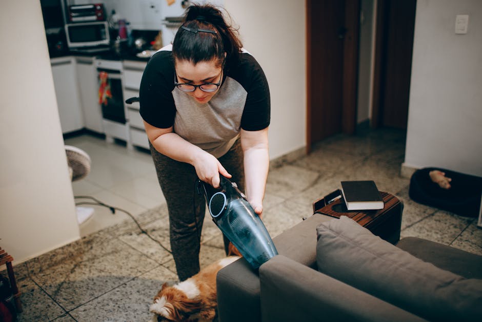 A person kneeling on a patterned carpeted floor, using a yellow and black vacuum cleaner to perform surface cleaning. The individual is holding a white cloth to clean the vacuum's dust compartment or 