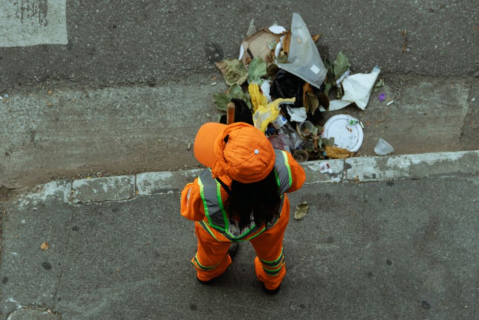 A person dressed in a grey uniform is performing surface cleaning in a modern indoor setting, possibly a waiting area or auditorium. They are using a handheld vacuum cleaner and a dustpan, suggesting 